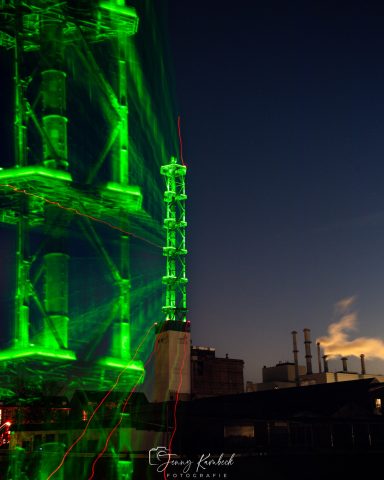 Grünes Licht strahlt von einem Fahrgeschäft bei Dämmerung, im Hintergrund Industrieanlagen.