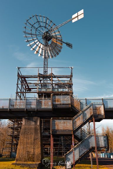 Windrad auf einer hohen Konstruktion mit Treppe und blauem Himmel im Hintergrund.