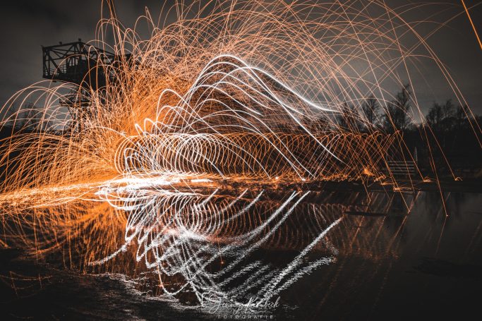 Lichtspuren in orange und weiß auf schwarzem Hintergrund, reflektiert im Wasser.
