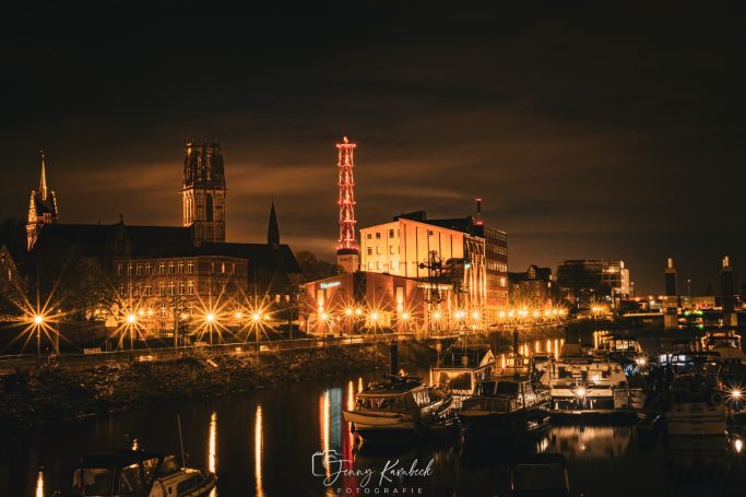 Nachtansicht einer Stadt mit beleuchteten Gebäuden und einem Fluss im Vordergrund.