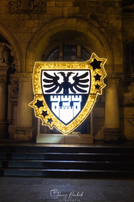 Leuchtendes Wappen mit Adler und Stadtmauer vor historischer Architektur.