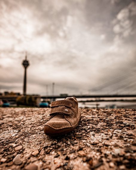 Ein einzelner Schuh liegt auf einem Kiesweg mit einem Fluss und einem Turm im Hintergrund.