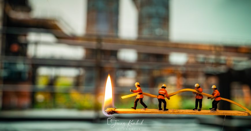 Miniaturfiguren von Feuerwehrleuten gehen eine brennende Zündholzschachtel entlang.