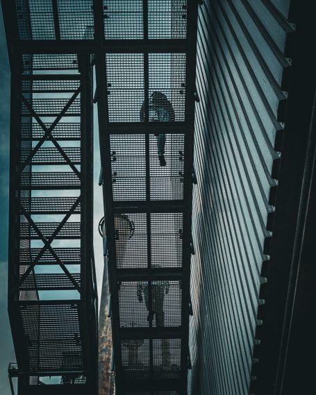 Zwei Personen stehen auf einer Feuerleiter zwischen Gebäuden, mit dramatischer Beleuchtung.
