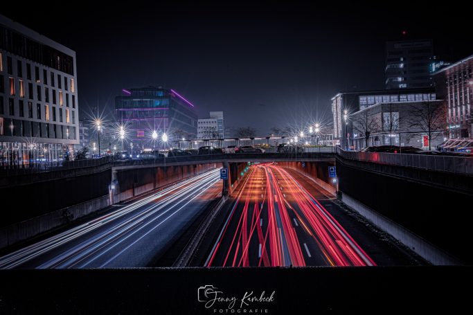 Lichtspuren von Autos über eine Stadtbrücke bei Nacht, umgeben von beleuchteten Gebäuden.