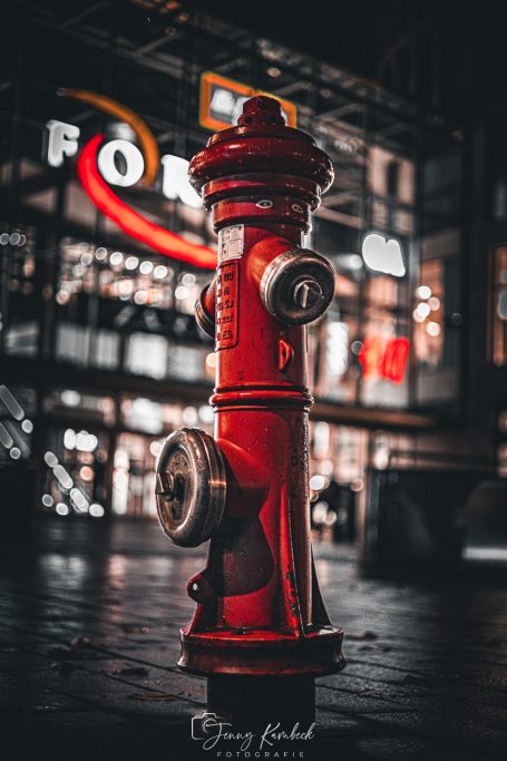 Rote Feuerwehrhydrant vor einem beleuchteten Hintergrund in der Nacht.