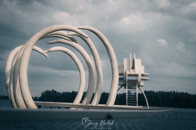 Abstrakte Skulpturen vor einem bewölkten Himmel am Wasser.