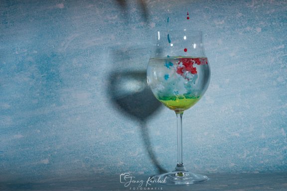 Ein Glas mit Wasser, gefüllt mit bunten Tropfen, vor einem blauen Hintergrund.