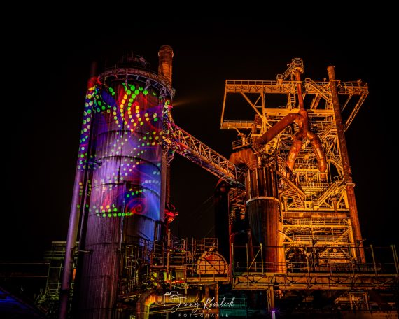 Bunt beleuchtete Industrieanlage mit großen Rohren und Metallstrukturen bei Nacht.