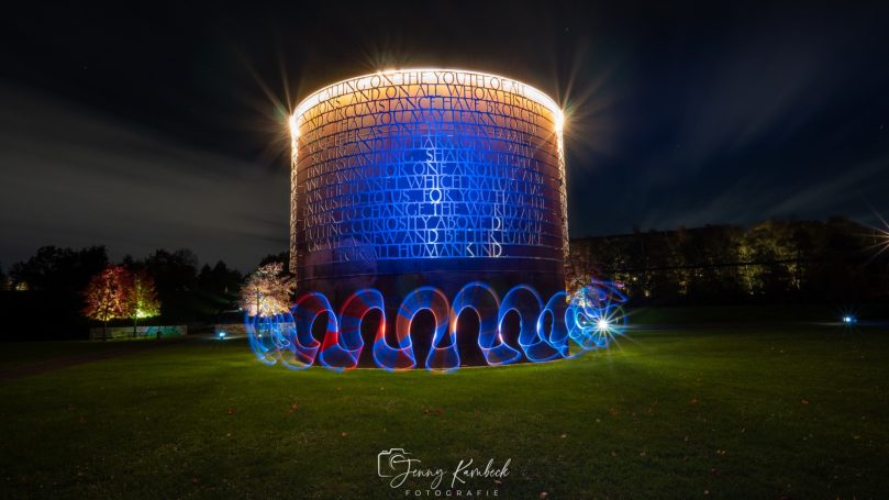 Bunt beleuchteter Wassertank bei Nacht, umgeben von grünem Rasen und Bäumen.